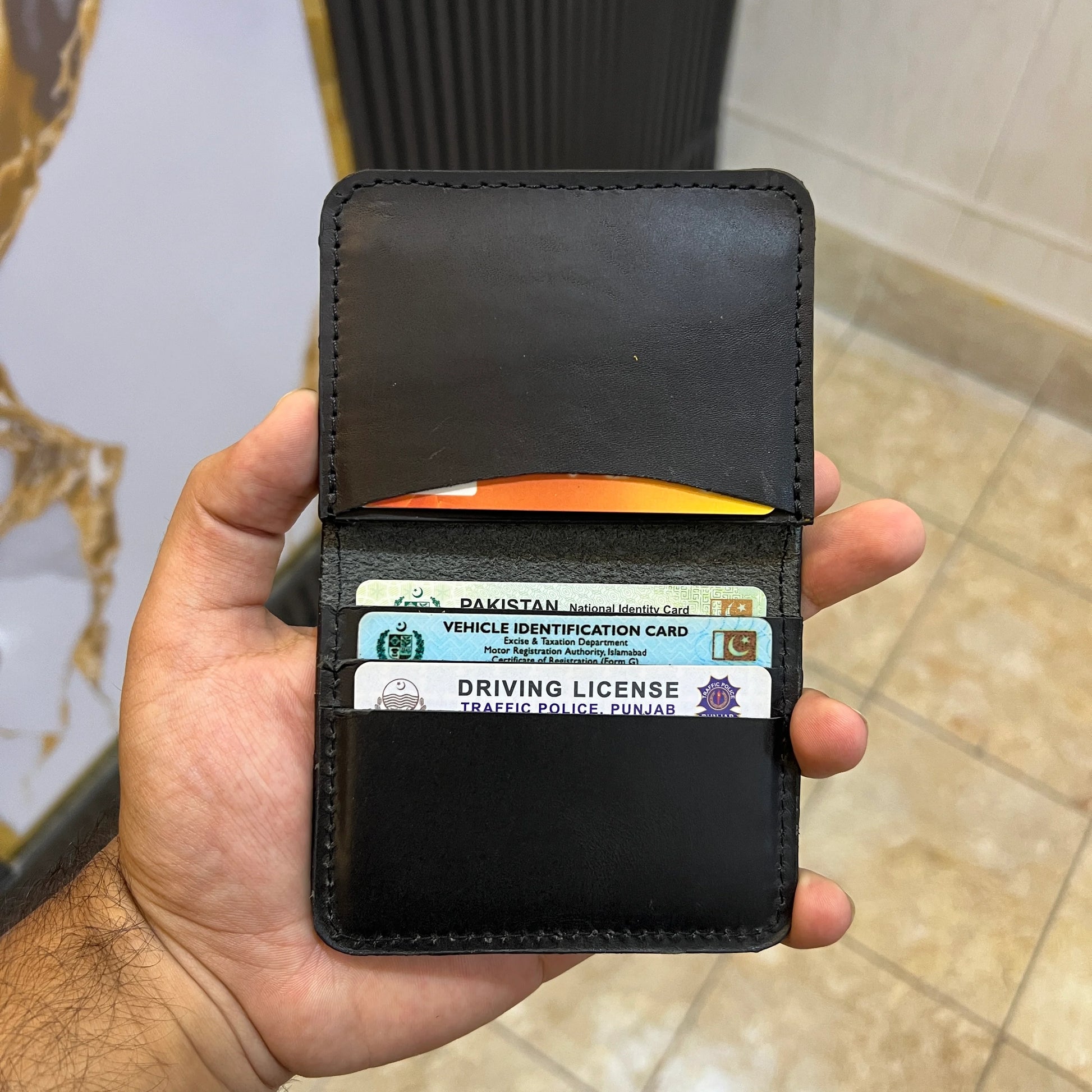 Hand holding a black wallet with cards, including a Pakistan vehicle identification card and driving license, against a blurred background.